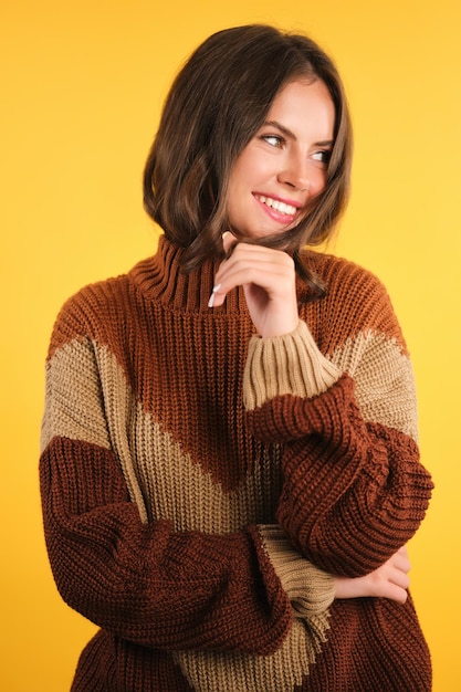 Beautiful smiling girl in cozy sweater joyfully looking aside over yellow background