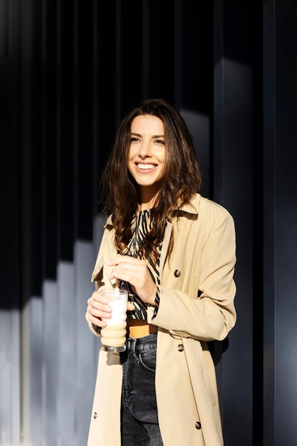 Beautiful smiling girl holds a glass in her hands stands on the background of a gray wall on the street Attractive girl in a casual beige coat is drinking a drink outdoors