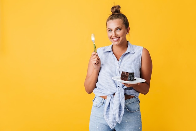 Beautiful smiling girl in shirt happily looking in camera while holding plate with slice of chocolate cake over yellow background