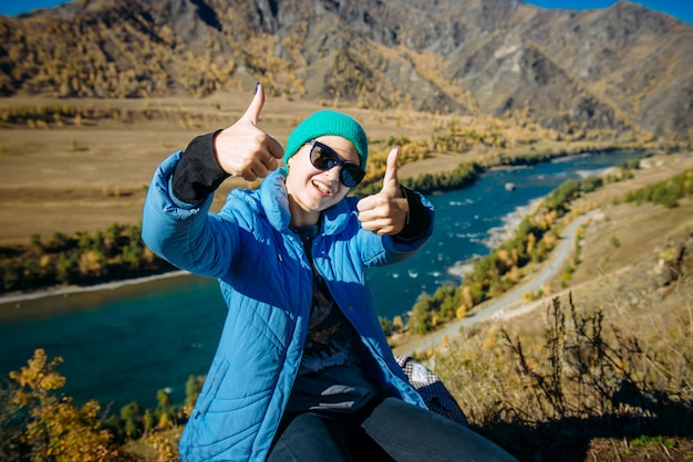 Beautiful smiling girl in sunglasses shows thumbs up, sitting on hill, enjoying autumn day in the Altay mountains.