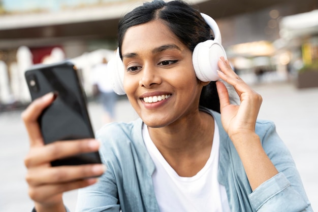 Beautiful smiling Indian woman holding mobile phone listening music in wireless headphones