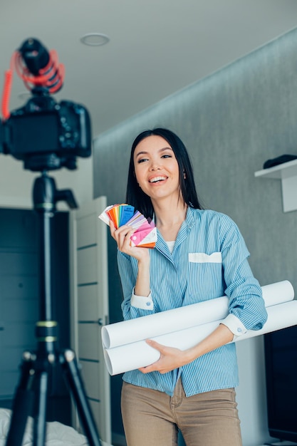 Beautiful smiling lady with blueprints and color palette samples recording a video and looking at the camera on tripod