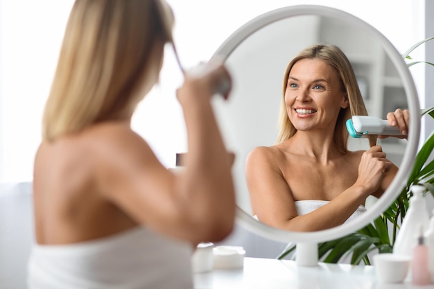 Beautiful smiling mature woman using hair straightener at home