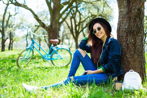 Beautiful smiling stylish hipster girl in a hat and sunglasses is sitting in a park under a tree with a phone in her hands and listening to music on a retro bike background