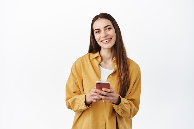 Beautiful smiling woman making order online shopping in application holding smartphone and looking happy at camera standing over white background