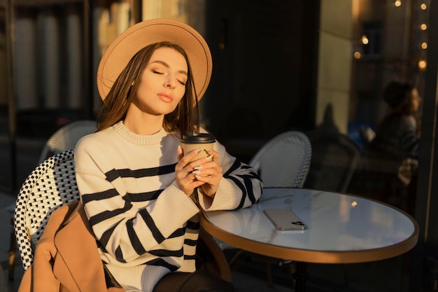 Beautiful smiling woman in stylish outfit sitting at the table in a hat and sweater romantic happy mood waiting for a boyfriend on a date in a cafe springsummer fashion trend drinking coffee