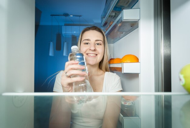 Beautiful smiling woman taking water from fridge and drinking it. View from inside of open refrigerator