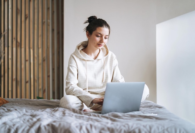 Beautiful smiling woman teenager girl student freelancer with dark long hair in casual using laptop sitting on bed at the home