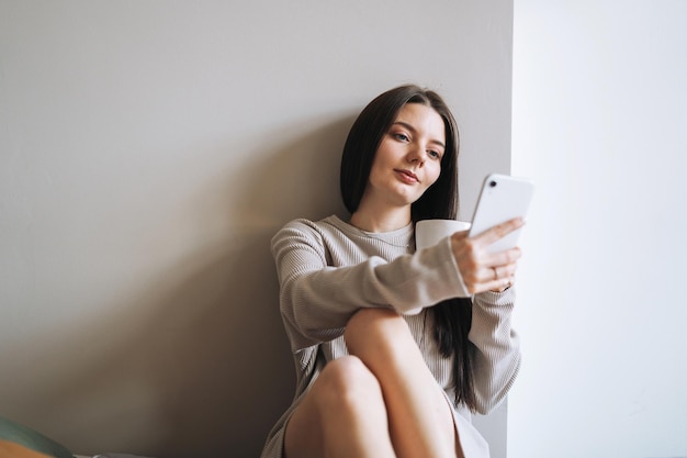 Beautiful smiling woman teenager girl student with dark long hair in home clothes with morning cup of coffee or tea using mobile phone cosial media on the grey background