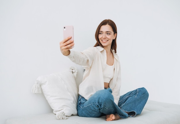 Beautiful smiling woman teenager girl student with dark long hair in white shirt taking selfie on mobile phone in hand sitting on couch at home