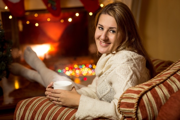 Beautiful smiling woman in white sweater sitting at fireplace