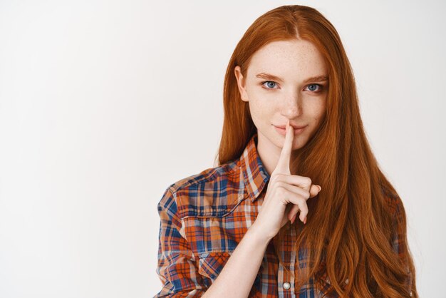 Beautiful smiling woman with long ginger hair and blue eyes sharing a secret hushing with finger on lips standing over white background