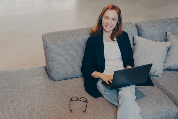 Beautiful smiling woman with red hair sitting with laptop and wireless headset on sofa in office