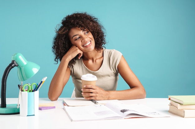 Beautiful smiling young african businesswoman casually dressed sitting at the desk isolated over blue wall, holding takeaway coffee cup