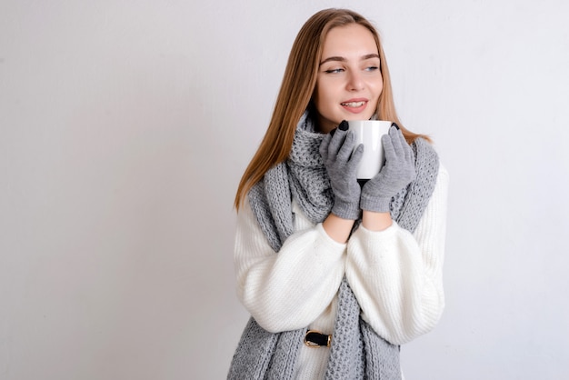 Photo beautiful smiling young blonde woman in white sweater, scarf and gloves holding a white cup of drink in her hands.