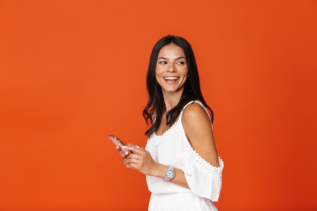 Beautiful smiling young brunette girl wearing white dress standing isolated over red wall, using mobile phone