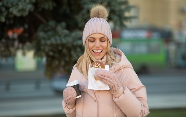 Beautiful smiling young woman in warm clothing with cup of hot tea coffee or punch. Woman eating cookies outdoor.