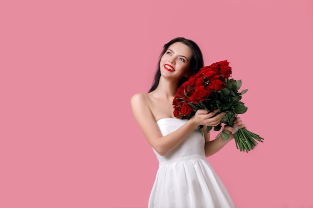 Beautiful smiling young woman in a white dress with a large bouquet of red roses on pink background