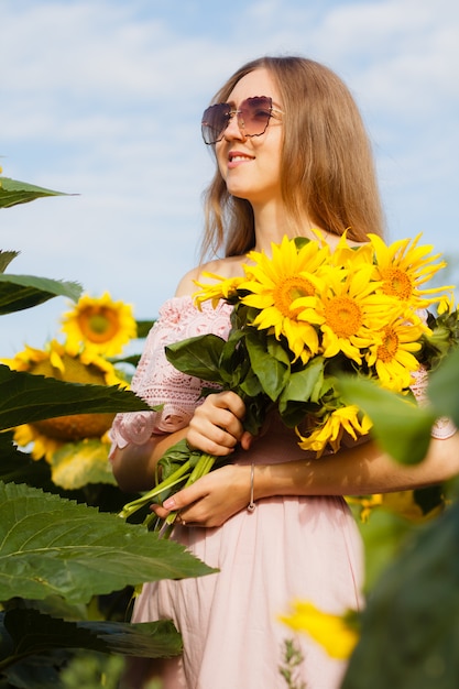 Beautiful smiling young woman in a white shirt stands in the field among sunflowers