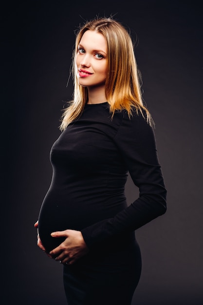 Beautiful soft and sensual pregnant woman in black dress standing against a background of gray wall in the studio.