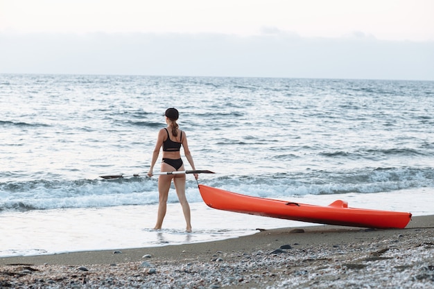 Beautiful sports girl in a black swimsuit goes with an orange kayak to swim in the sea at sunset