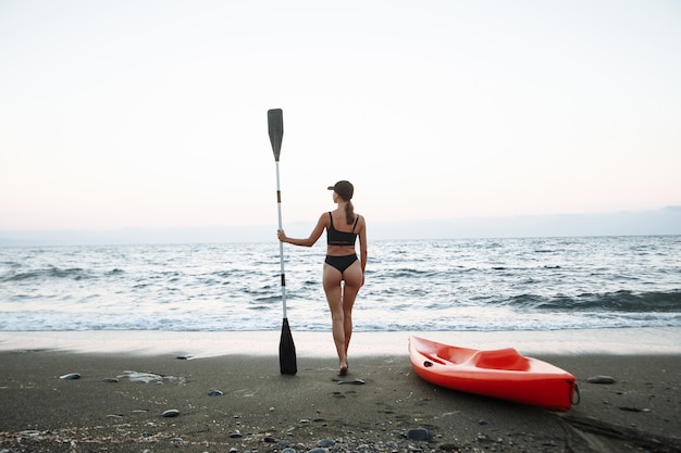 Beautiful sports girl in a black swimsuit goes with an orange kayak to swim in the sea at sunset