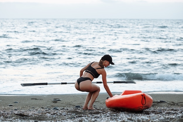 Beautiful sports girl in a black swimsuit goes with an orange kayak to swim in the sea at sunset