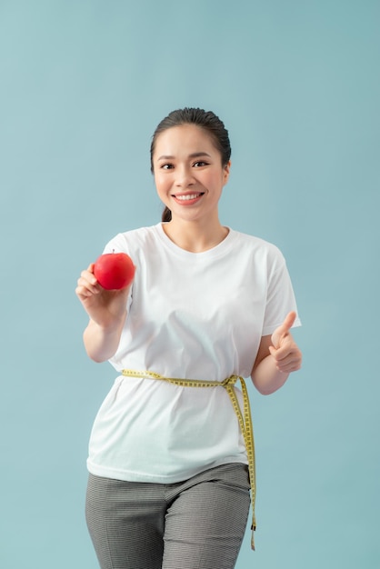 Beautiful sporty woman using tape measure eating apple fruit showing thumb up smiling happy with open mouth