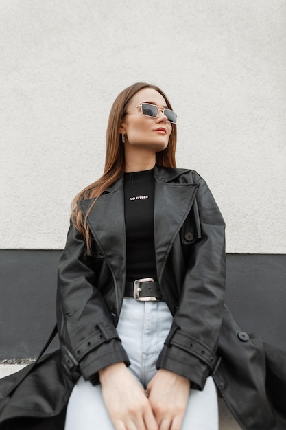 Beautiful street fashion girl in fashionable clothes with vintage gold sunglasses in a black leather coat with a black Tshirt and jeans sits in the city near the building