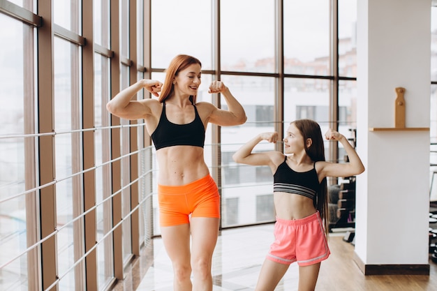 Beautiful strong woman and charming little daughter are showing their biceps and smiling while working out at gym