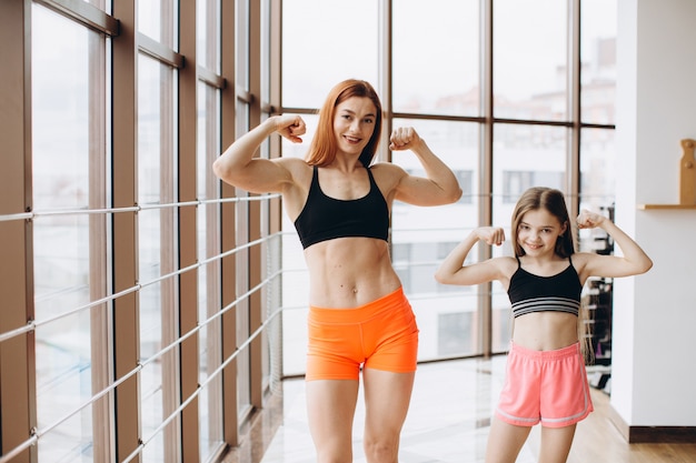 Beautiful strong woman and charming little daughter are showing their biceps and smiling while working out at gym