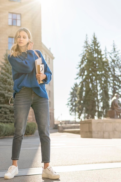 Beautiful student with notebooks in hand straightens her hair, against the background of the university building on a sunny day