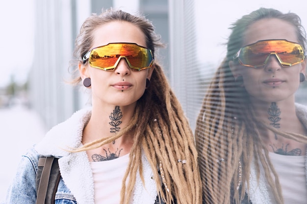 Photo beautiful styilish girl in big toned glasses, tattoo on the neck and piercing is posing near mirroed wall.