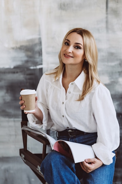 Beautiful stylish blonde woman in denim casual clothes and a white blouse with voluminous sleeves with a magazine and coffee in her hands. Soft selective focus.