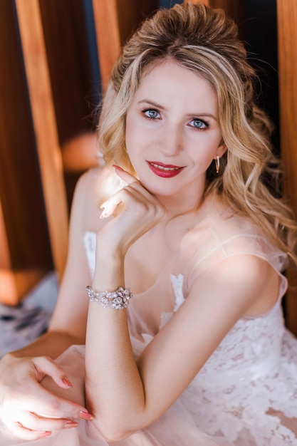 A beautiful stylish bride in a long light pink wedding dress poses in a bright hotel lobby sitting on a carpet near a wooden wall Bride portrait