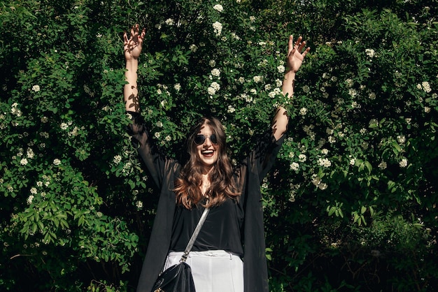 Beautiful Stylish Hipster Girl with beautiful hair having funfalling in floral bushes in sunny day Boho Woman with sunglasses in fashionable outfit smiling and enjoying day in garden