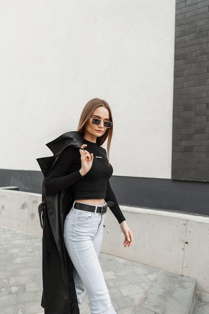 Beautiful stylish urban model woman with gold vintage eyewear in fashionable urban casual clothes with black Tshirt and high waisted jeans with coat walking on the street near the building