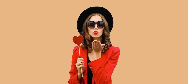 Beautiful stylish woman blowing kiss with sweet red heart shaped lollipop on stick in black hat