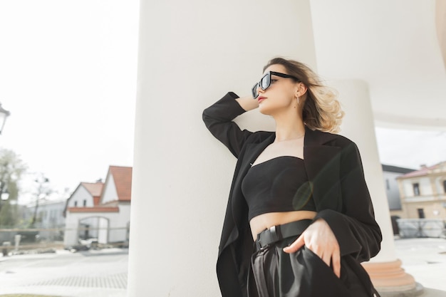 Beautiful stylish young girl hipster model with sunglasses wearing a fashion black strict clothes with a jacket top and pants is standing near a white column in the city