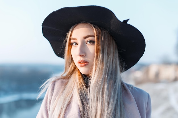 Beautiful stylish youth woman model with a black hat in a fashion coat on a sunny winter day outdoors Pretty lady