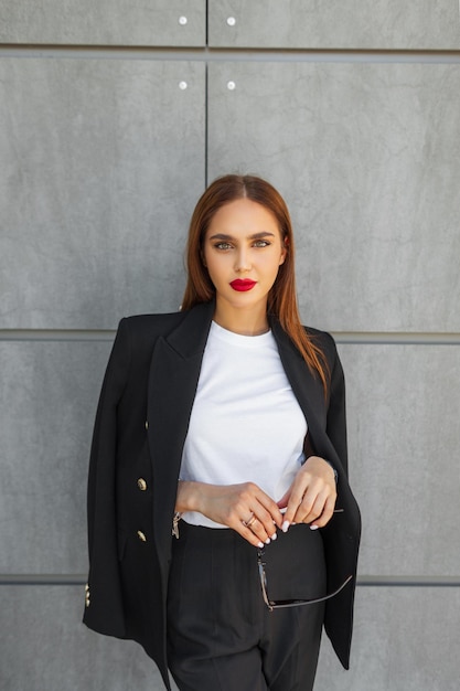 Photo beautiful successful young stylish business woman model with sunglasses in fashion casual clothes with a white tshirt and blazer stands near a gray wall in the city