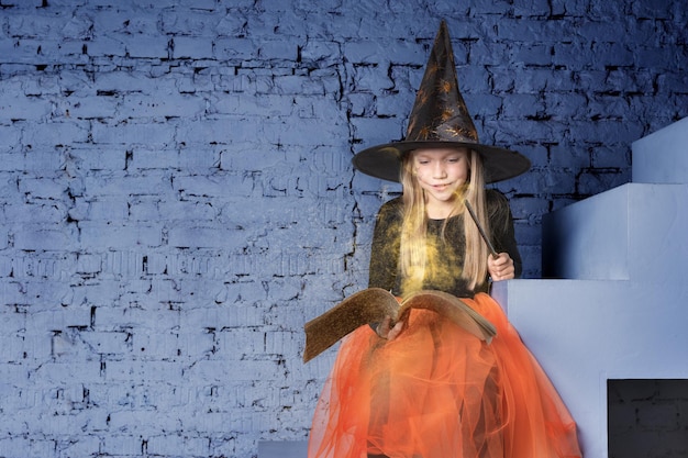 A beautiful sweet girl in a witch costume wearing a hat holding a book with spells and a magic wand conjures