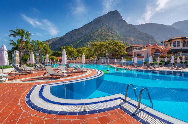 Beautiful swimming pool with sun beds and umbrellas at sunrise in summer Luxury resort Liberty hotels lykia Oludeniz Turkey Landscape with empty pool deck chairs green trees mountain blue sky