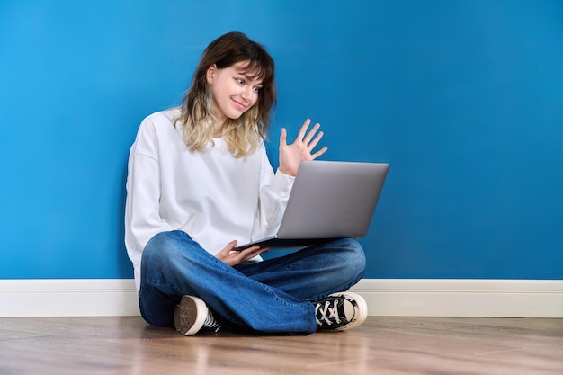 Beautiful teenage female sitting on the floor with laptop on blue background