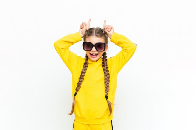 A beautiful teenage girl shows horns or points at your advertisement on a white isolated background A child in a yellow summer suit and sunglasses