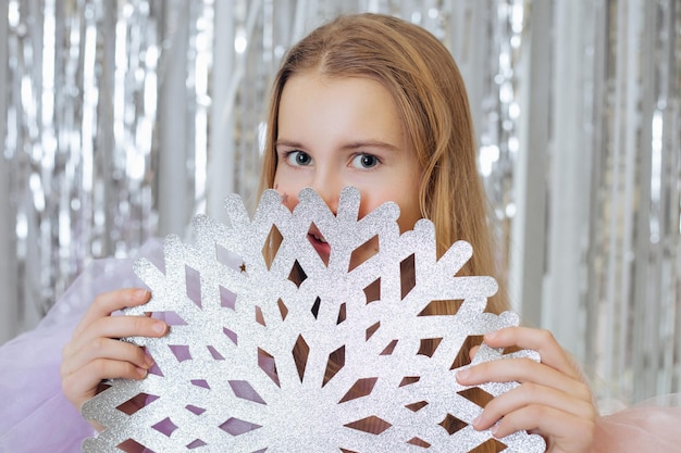 Beautiful teenage girl smiles mysteriously hiding behind large paper snowflake which she holds in her hands New Year and Christmas Decoration and decor Happy emotions Festive mood