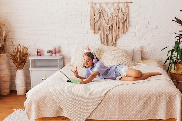 Beautiful teenager girl communicates online by phone while lying on the bed at home. Child in headphones learn online while lying on the bed. Distance learning concept