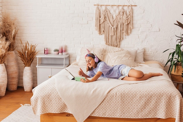 Beautiful teenager girl communicates online by phone while lying on the bed at home. Child in headphones learn online while lying on the bed. Distance learning concept