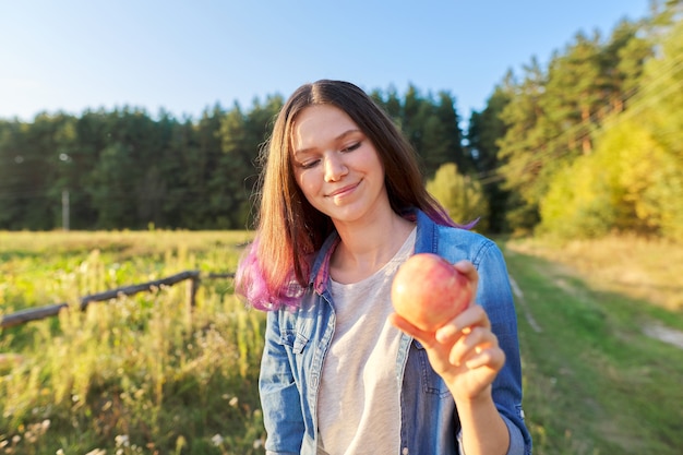 Beautiful teenager girl with red apple, happy young woman on nature, scenic sunset landscape background, copy space, healthy natural food concept