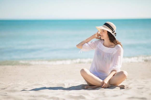 Beautiful teenager teen girl sitting on the beach wearing white beachwear and straw hat Summer holidays and vacation concept